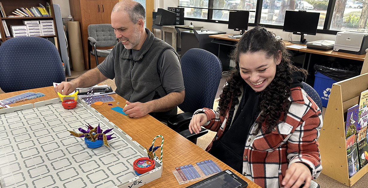 Pre Service Teacher Training 1230px Two educators smiling at a table while working with small robots on a grid with tablets.